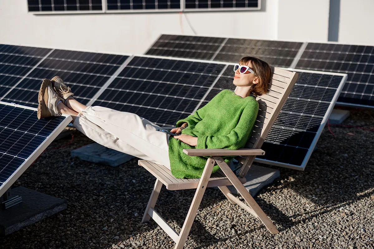 eine Frau sitzt in einem Liegestuhl vor mehreren Solarmodulen auf einem Dach und legt die Füße auf einem Modul hoch